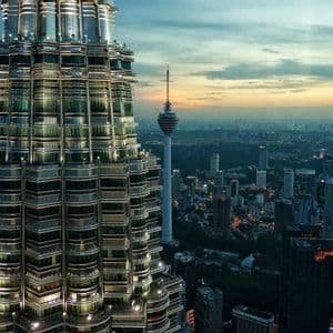 Un grattacielo illuminato domina un vasto paesaggio urbano con un'altra alta torre in lontananza al tramonto.