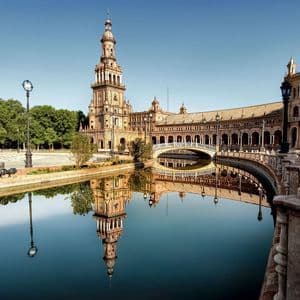 Un edificio storico ornato e la sua torre che si riflettono nell'acqua immobile di un canale sotto un cielo azzurro limpido.