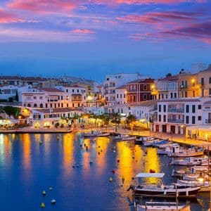 A coastal town's harbor illuminated at dusk, with lights from buildings reflecting on the water where boats are moored.