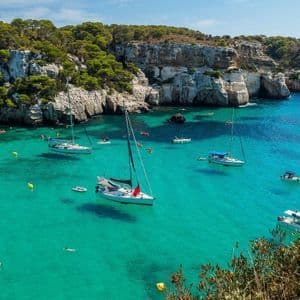 An aerial view of sailboats anchored in a calm, turquoise cove, framed by rocky cliffs topped with green trees.