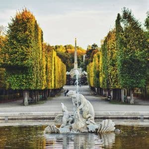 Una fontana di pietra in uno specchio d'acqua, con un lungo viale alberato da alti alberi autunnali che conduce a un monumento in lontananza.