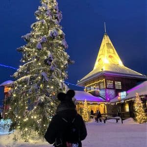 Eine Person mit WeRoad Rucksack betrachtet nachts auf einem Dorfplatz einen großen, schneebedeckten und beleuchteten Weihnachtsbaum.