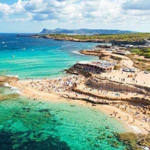 Veduta aerea di una caletta affollata con persone che nuotano e prendono il sole in acque limpide e turchesi vicino a una costa rocciosa in una giornata di sole.