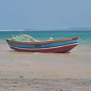 Una barca da pesca variopinta, di nome Inora, è ormeggiata in acque basse e turchesi su una spiaggia sabbiosa sotto un cielo azzurro e limpido.