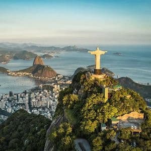 Vista aerea del Cristo Redentore su una vetta montuosa, con vista su una città costiera, una baia e altre montagne all'alba.
