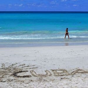 La palabra 'CUBA' y un sol están dibujados en la arena blanca de una playa mientras una persona camina en el océano turquesa.
