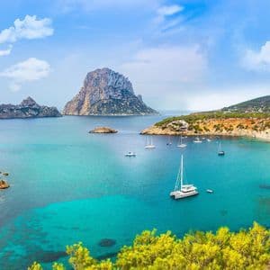 Barche a vela e un catamarano sono ancorati in una baia calma e turchese, circondata da scogliere rocciose e una grande isola.