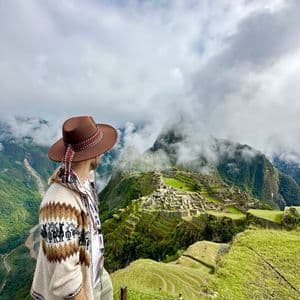 Eine Person in gemusterter Strickjacke und braunem Hut steht auf einem grasbewachsenen Hügel mit Blick auf antike Ruinen, eingebettet in neblige grüne Berge.