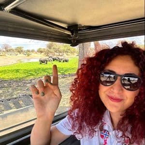 Una donna con capelli rossi ricci e occhiali da sole si scatta un selfie all'interno di un veicolo safari, con una mandria di elefanti visibile sullo sfondo.