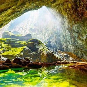 Sunlight streams through a large opening into a cavern, illuminating moss-covered rocks and a reflective green pool of water.