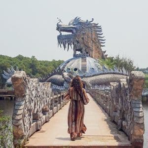 A woman seen from behind walks on a stone bridge towards a large, weathered sculpture of a dragon built over a lake.
