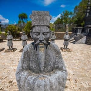 Una statua in pietra consumata di un uomo in abiti tradizionali si erge in un cortile pavimentato con altre statue sullo sfondo.