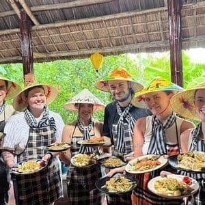 A WeRoad group trip of six people wearing conical hats and aprons, smiling and holding plates of food at a cooking class.