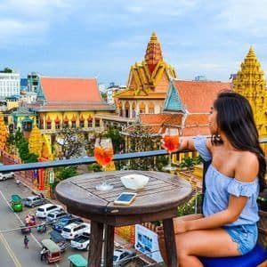 A woman sits at a table on a rooftop balcony with two cocktails, overlooking a city with golden temples and street traffic.
