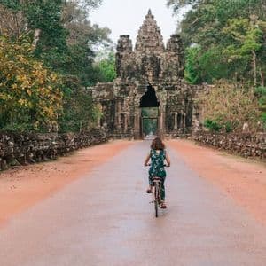 Una persona vista di spalle pedala in bicicletta su una strada che porta a un grande, antico portale in pietra affiancato da alberi della giungla.