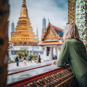 A woman with blonde hair in a green shirt leans on an ornate balcony, looking out at a golden temple.