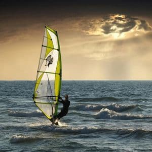 Eine Person windsurft bei Sonnenuntergang auf dem Meer mit einem lindgrünen Segel unter bewölktem Himmel.