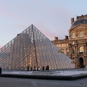 A large glass pyramid stands in a paved courtyard in front of an ornate, historic building under a pink and blue sky at dusk.