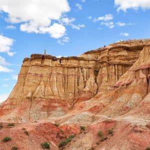 Une personne se tient au bord d'une formation rocheuse imposante, stratifiée, de couleurs rouge et jaune, sous un ciel bleu partiellement nuageux.