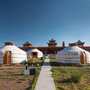 Plusieurs yourtes blanches sont installées dans un champ verdoyant, devant un grand bâtiment de style traditionnel, sous un ciel bleu dégagé.