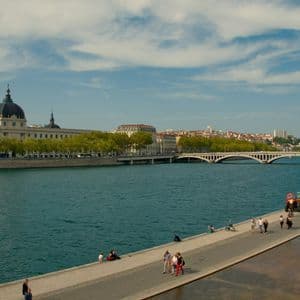 La gente passeggia e si rilassa su un lungofiume in una città, con un edificio a cupola e un grande ponte sullo sfondo.