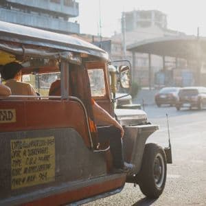 Un Jeepney rosso e argento trasporta passeggeri lungo una soleggiata strada cittadina, con edifici e altre auto sullo sfondo.