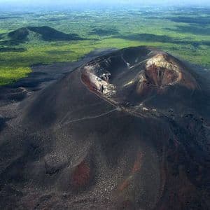 Una vista aerea di un vulcano scuro con escursionisti sul pendio che si dirigono verso il cratere, immerso in un lussureggiante paesaggio verde.