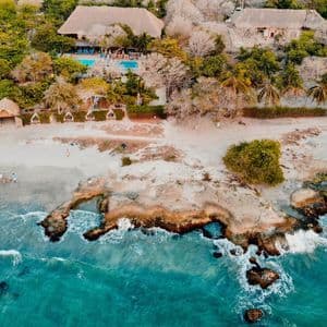 Una vista aérea de una costa tropical donde el agua turquesa se encuentra con una playa de arena y un resort con edificios de techos de paja entre árboles.