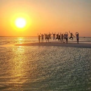 A WeRoad group trip is silhouetted against a golden sunset, jumping together on a sandbar surrounded by the sea.