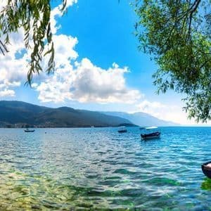 Ein kleines Boot liegt an einem klaren, ruhigen See mit entfernten Bergen, umrahmt von grünen Ästen unter blauem Himmel.