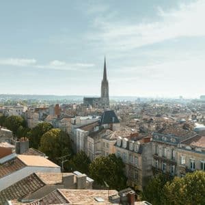 Una vista aérea de una ciudad europea con tejados de terracota, con un alto campanario de iglesia elevándose en el centro bajo un cielo azul brumoso.