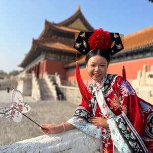 Una donna sorridente, in abito tradizionale rosso ricamato e copricapo, tiene in mano un ventaglio a forma di farfalla, con un palazzo cinese sullo sfondo.