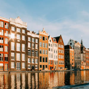 Une rangée de bâtiments colorés et traditionnels au bord d'un canal se reflétant dans l'eau sous un ciel clair et lumineux.