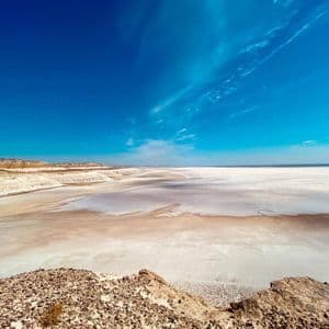 A panoramic view from a rocky outcrop over a vast, arid salt flat stretching to the horizon under a deep blue sky.