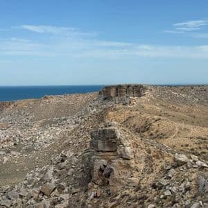 Una costa frastagliata e rocciosa, con scogliere a picco e vegetazione rada, si affaccia su un mare blu e tranquillo sotto un cielo sereno.