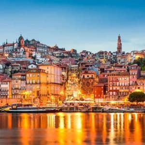 Un paesaggio urbano collinare colorato illuminato al crepuscolo, con le luci degli edifici che si riflettono sul fiume calmo sottostante.