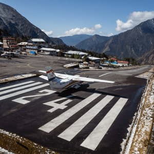 Un pequeño avión de hélice en una pista marcada con el número 24, situado en un valle de alta montaña con edificios cercanos.