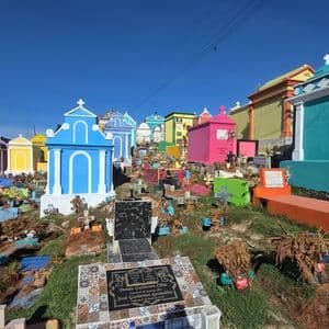 Un vibrante cementerio en una ladera con mausoleos de colores brillantes, en azul, rosa y amarillo, bajo un cielo azul claro.