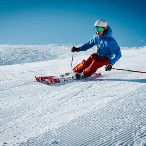 Una persona con giacca blu e pantaloni arancioni scia su una pista innevata battuta sotto un cielo azzurro limpido.