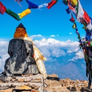 Una statua di pietra vista da dietro domina una catena montuosa innevata e un mare di nuvole, sotto colorate bandiere di preghiera tibetane.