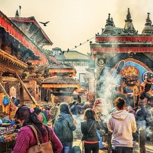 Una folla di persone si è radunata in una vivace piazza cittadina con architettura tradizionale, bancarelle e fumo che si alza davanti a un arco ornato.