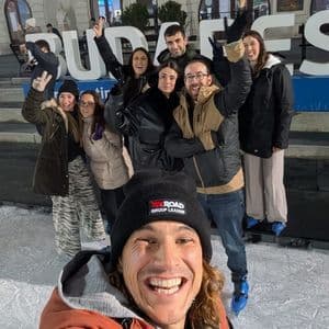 Un líder de grupo de WeRoad se toma una selfie sonriendo con su grupo mientras patinan sobre hielo al aire libre frente a un gran edificio.