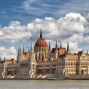 Un imponente e sontuoso edificio con una cupola rossa e alte guglie si erge sul bordo di un ampio fiume sotto un cielo azzurro nuvoloso.