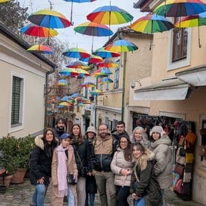 Un viaje en grupo WeRoad posa para una foto en una calle empedrada bajo un dosel de coloridos paraguas arcoíris.