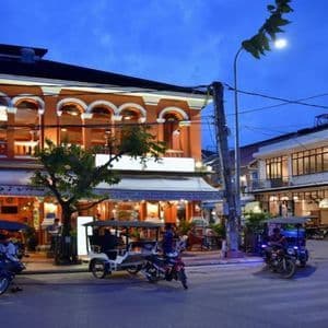 Tuk-tuks and scooters are parked on a city street corner in front of illuminated colonial-style buildings at dusk.
