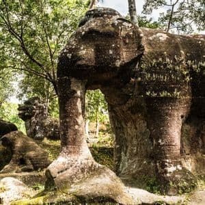 Una gran escultura de piedra cubierta de musgo de un elefante se alza entre estatuas de leones más pequeñas en un exuberante bosque verde.