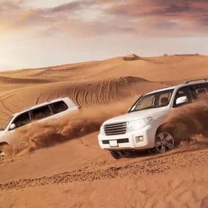 Deux SUV blancs traversent un désert, soulevant du sable alors qu'ils naviguent dans les grandes dunes sous un ciel de coucher de soleil brumeux.