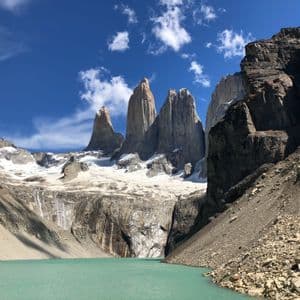 Three jagged granite peaks and a glacier towering over a turquoise glacial lake under a bright blue sky with sparse clouds.