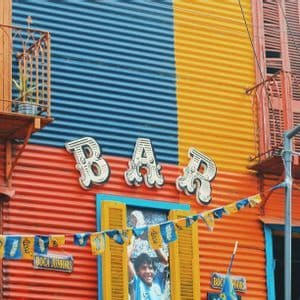 Una colorida fachada de edificio de metal corrugado en rojo, azul y amarillo, con un letrero de 'BAR' y pequeñas banderas colgadas.
