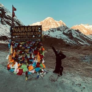 Eine Person macht einen Kopfstand neben dem Annapurna Base Camp Schild, mit sonnenbeschienenen, schneebedeckten Bergen im Hintergrund.
